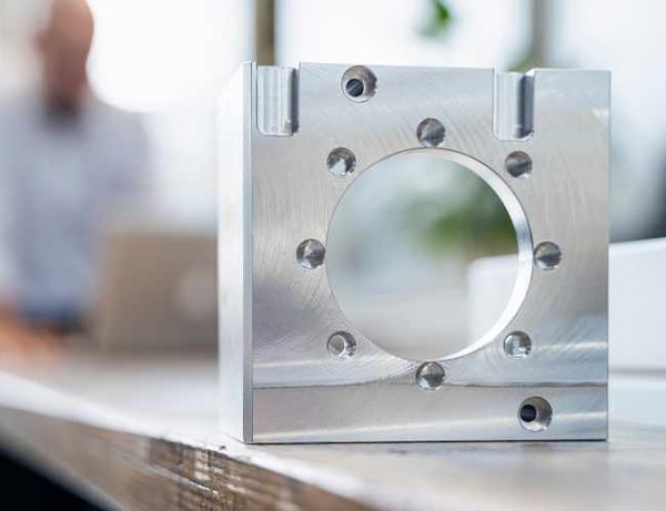 A finished aluminum component sits on a work bench with an engineer working in the background